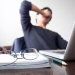 man with his hand holding his face taking a brake from working with laptop computer and notebook with eye glasses on wooden desk. concept of stress/rest/tension/failed/discourage